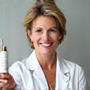 Μια έμπειρη δερματολόγος με λευκή ιατρική μπλούζα, δείχνει μια φιάλη ορού προσώπου με pipet σε ένα κομψό, δερματολογικό ιατρείο, με φόντο σύγχρονο εξοπλισμό και φυσικό φως.